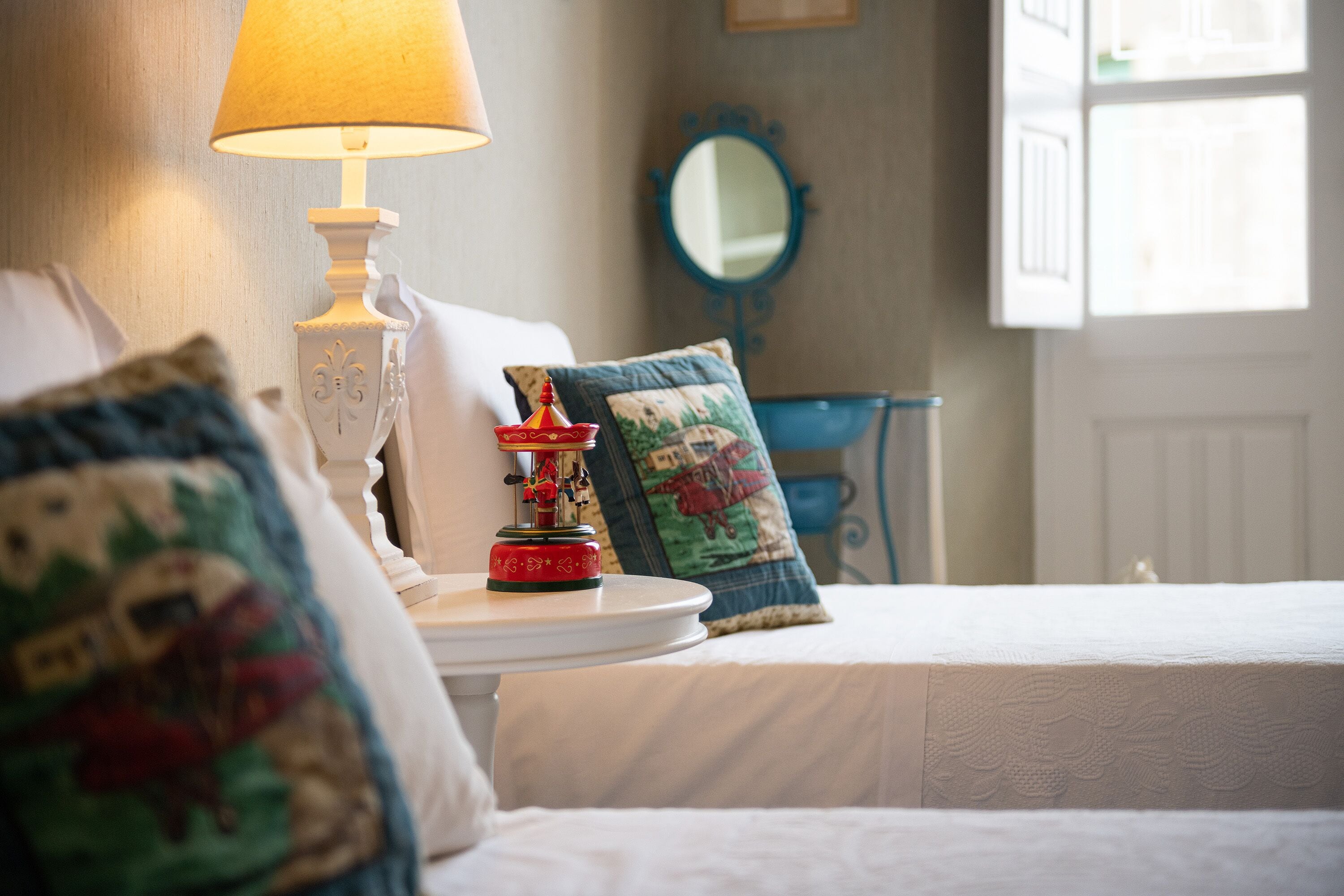 Un lit bien fait, avec une couette blanche et des coussins décoratifs, une petite table avec une lampe et un miroir au mur.