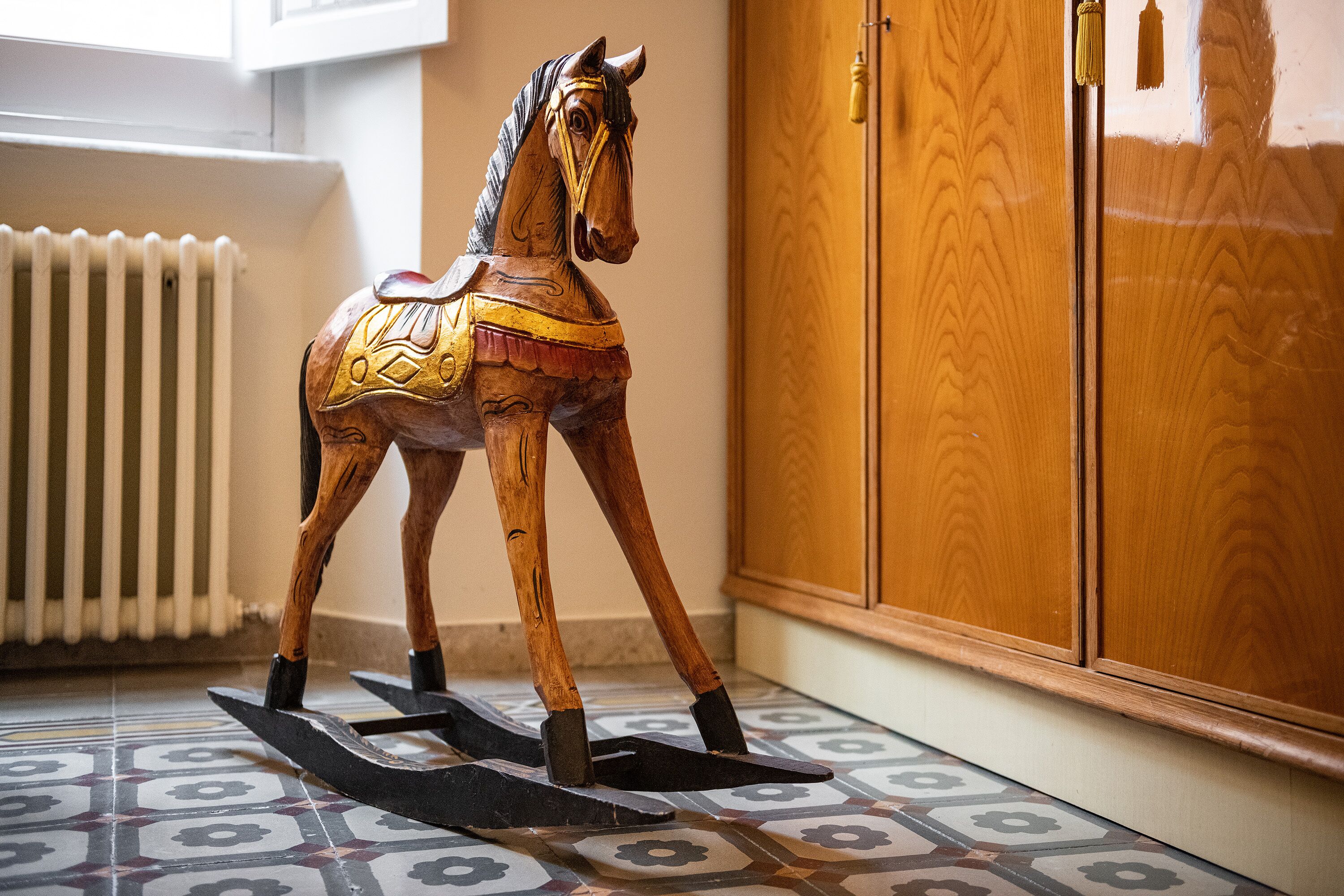 A wooden rocking horse in a room with patterned floor tiles and wooden cabinets.