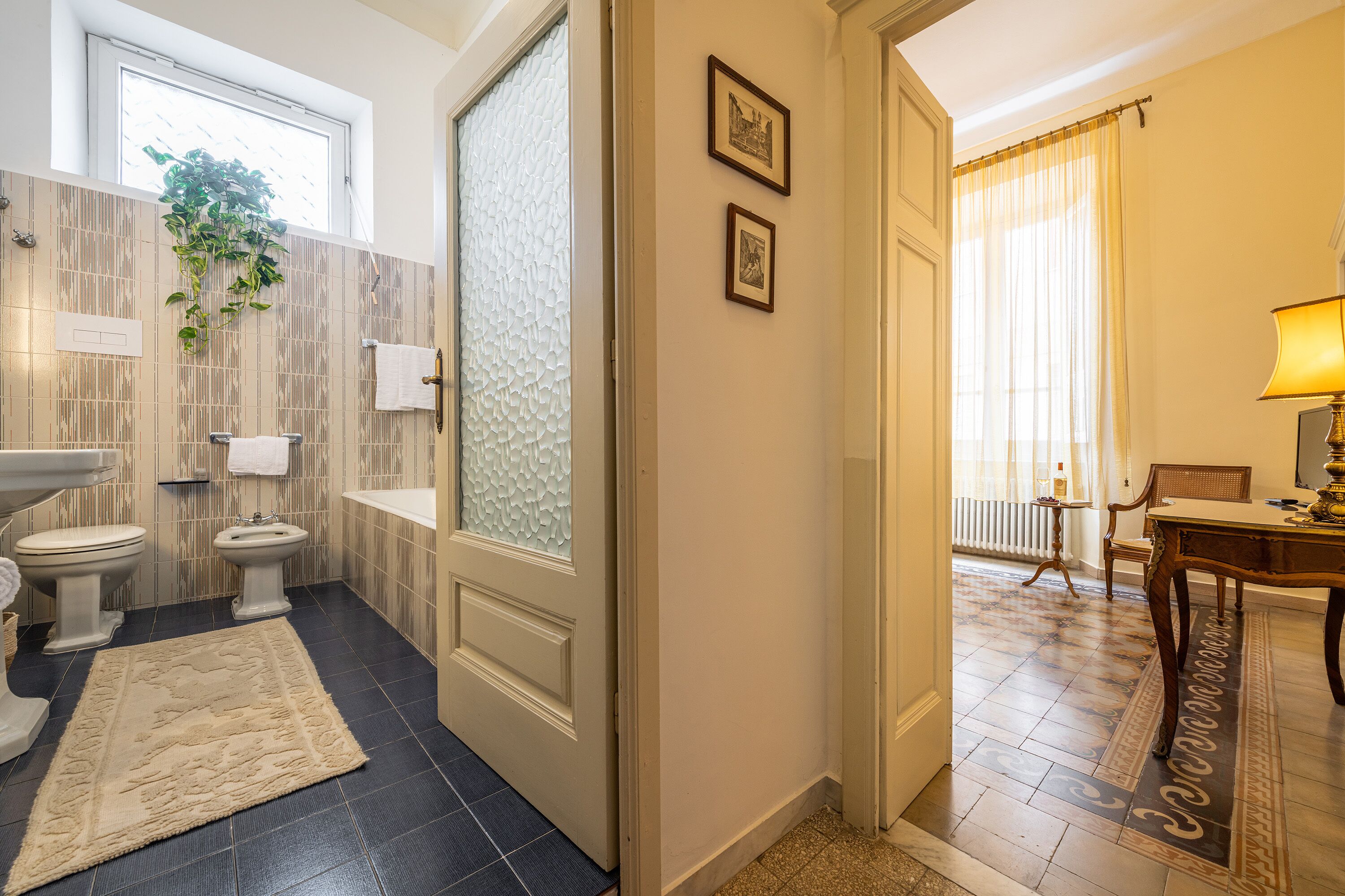 Ein Badezimmer mit Toilette, Bidet und Waschbecken. Auf dem Boden liegt ein Teppich und von der Decke hängt eine Pflanze.