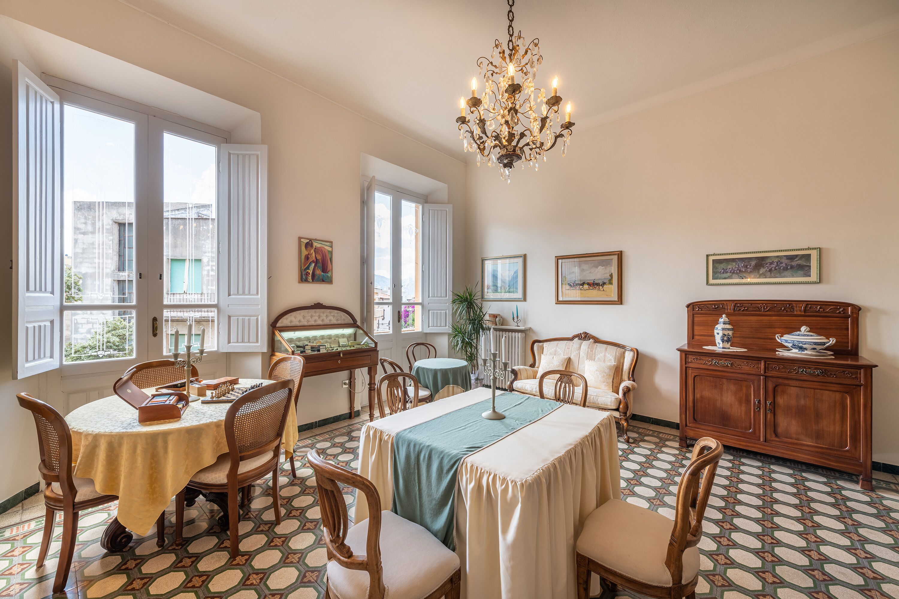 A dining area with a round table, chairs, a chandelier, and a view through large windows.