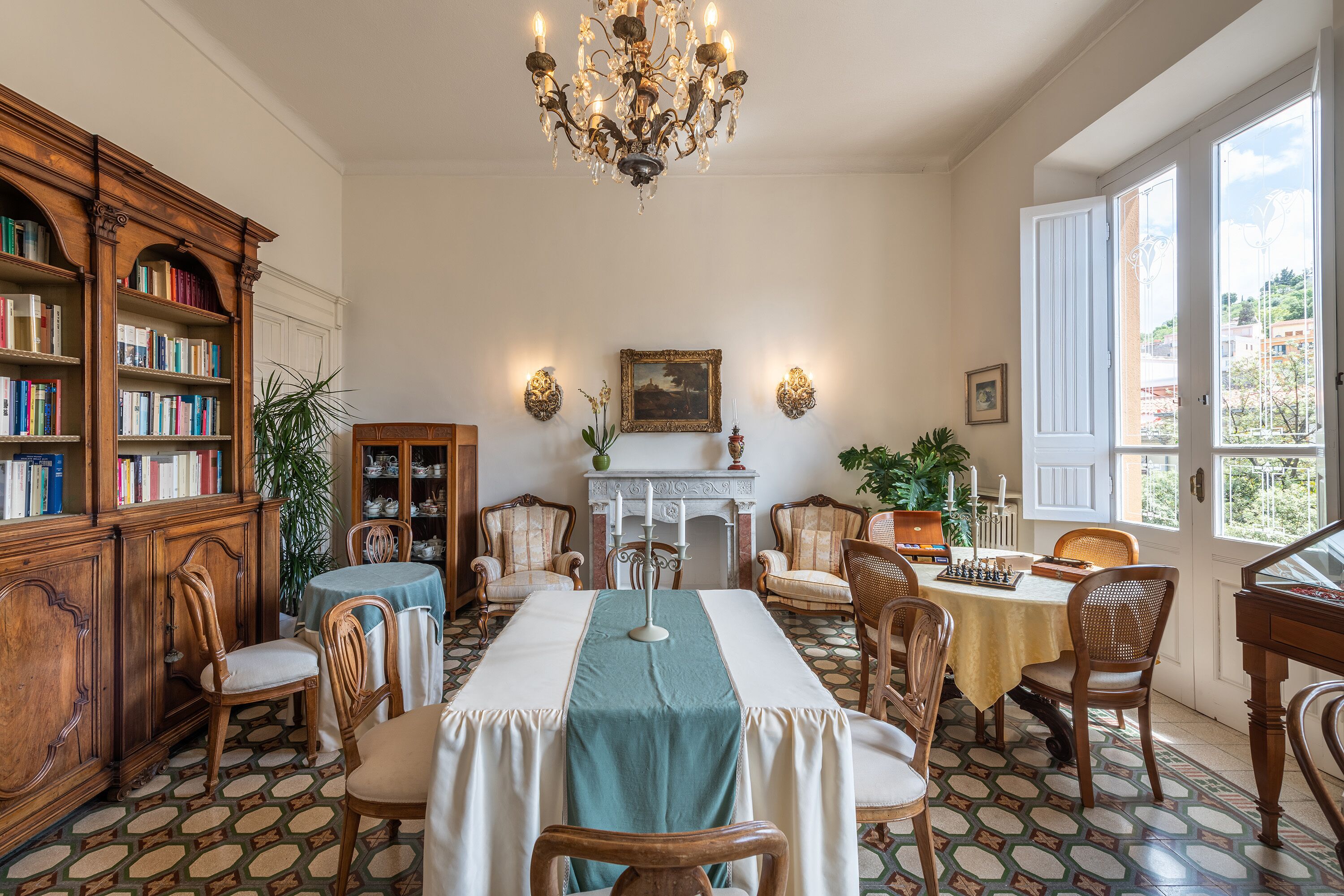 A dining area with a long table, chairs, a chandelier, bookshelves, and a fireplace.
