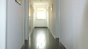 Un long couloir étroit, doté d’un sol carrelé foncé et de murs blancs. Des tableaux encadrés ornent les murs, et une fenêtre se trouve au bout du couloir.