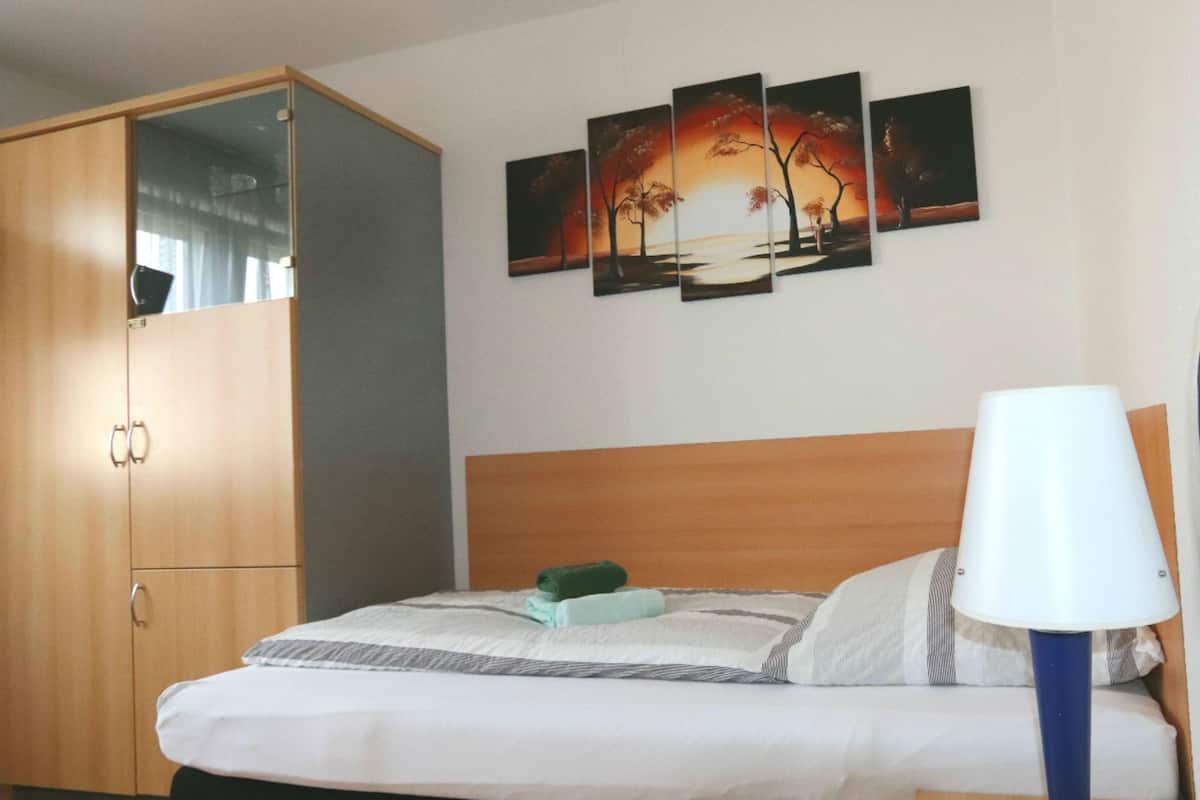 A hotel room with a bed, a wardrobe, a nightstand, a lamp, and a wall with three paintings.