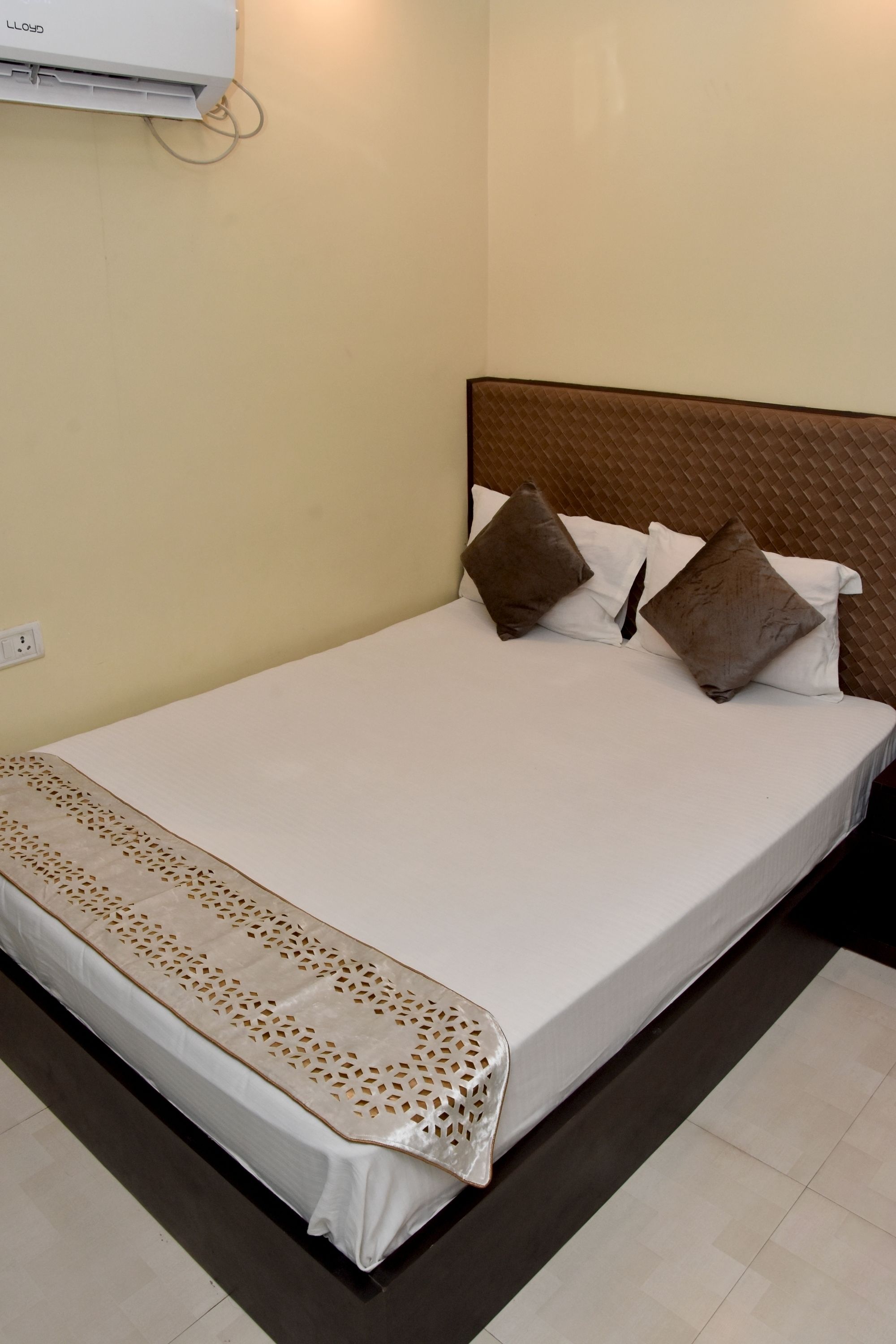 A neatly made bed with a white sheet and two brown pillows, a beige headboard, and a wall-mounted air conditioner.