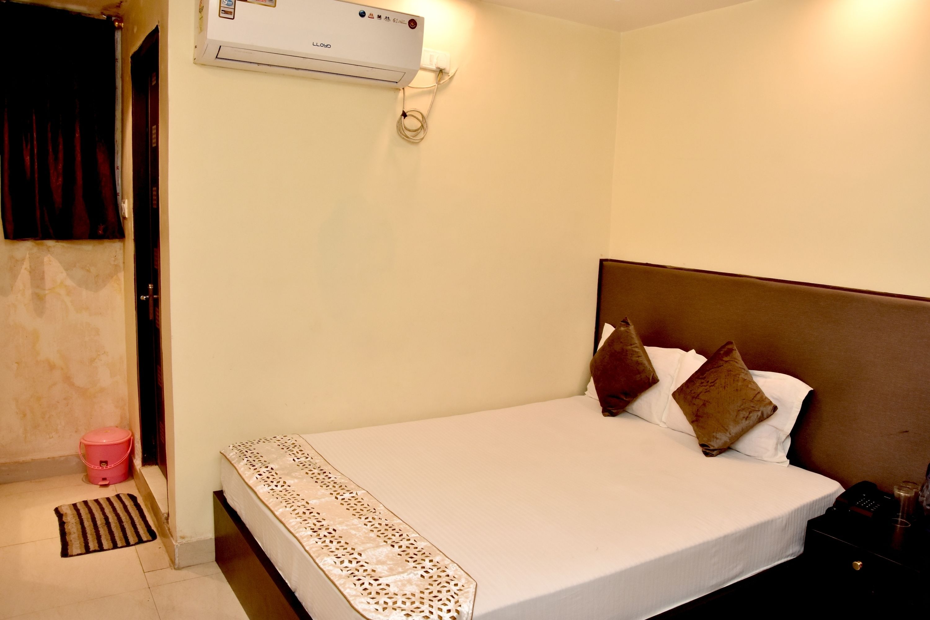 A hotel room with a bed, a bedside table, a lamp, and an air conditioning unit.