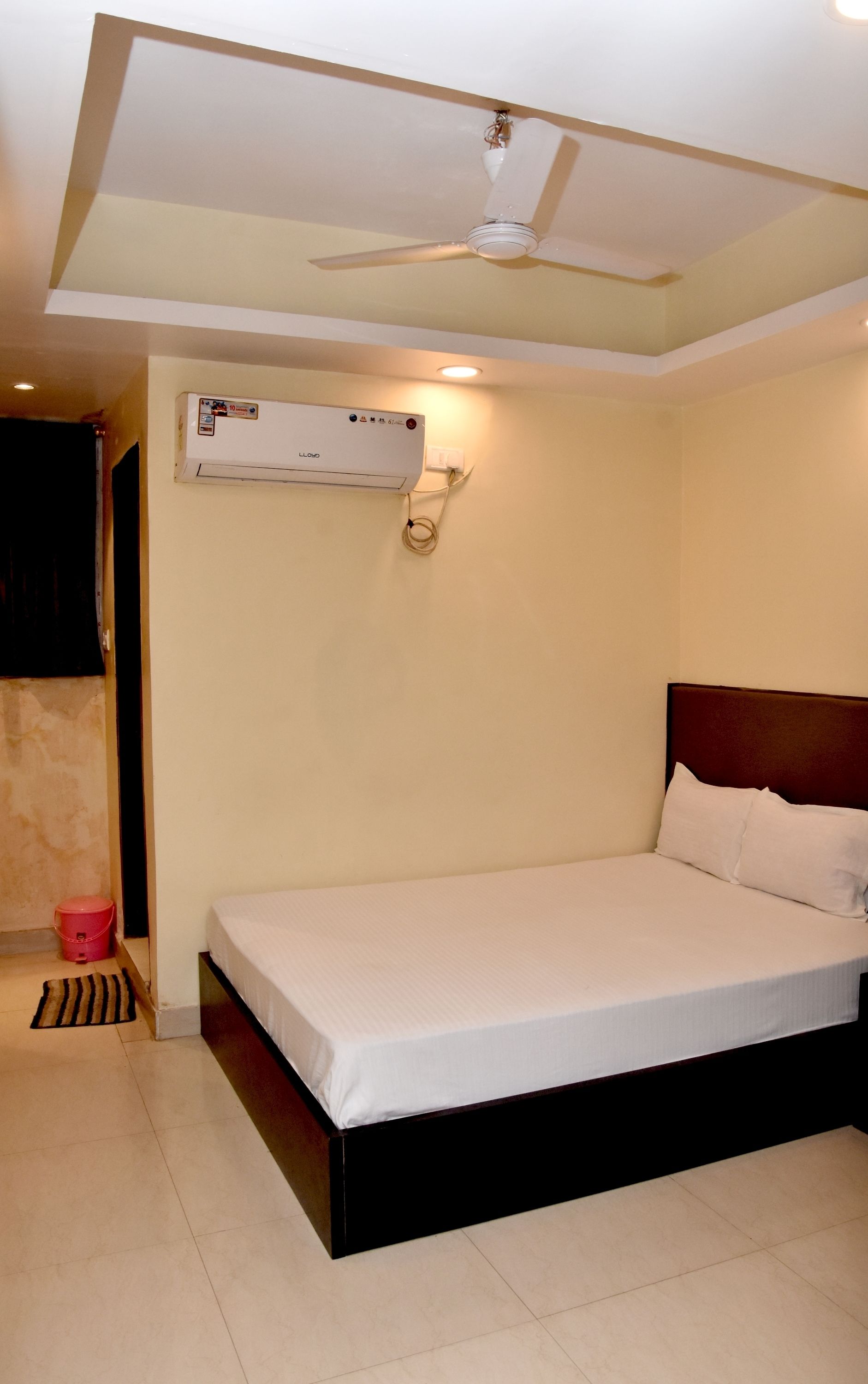 A bedroom with a bed, a ceiling fan, an air conditioning unit, and a red bucket on the floor.