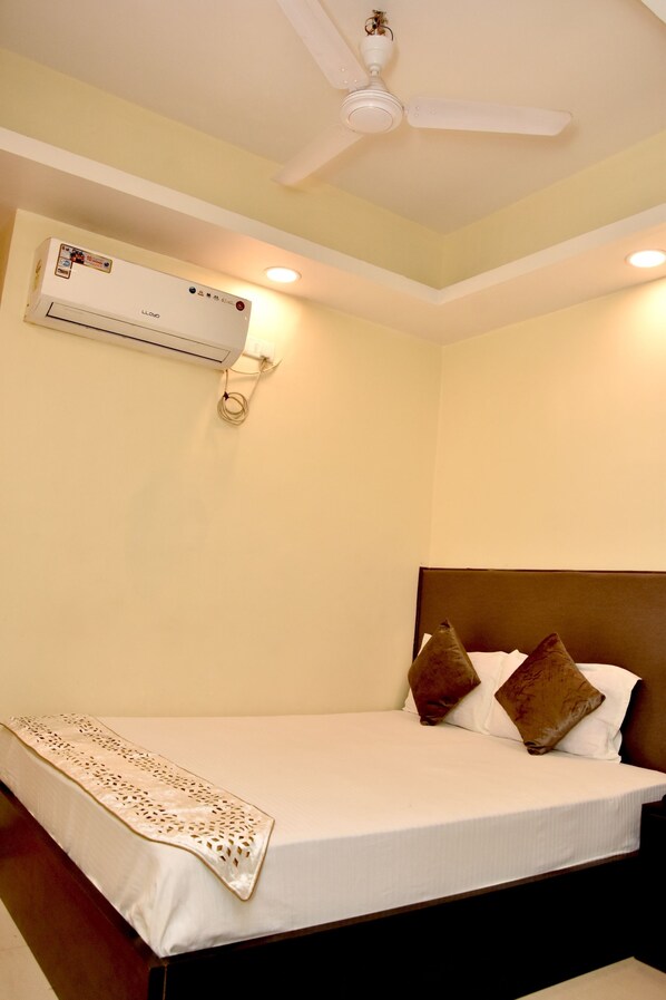 A neatly made bed with a white sheet and a decorative white lace throw, a ceiling fan, and an air conditioning unit mounted on the wall.