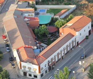 An aerial view of a building with a red-tiled roof, a swimming pool, and a street with parked cars. - Hostal Restaurante Cuatro Caminos (Piedrabuena)