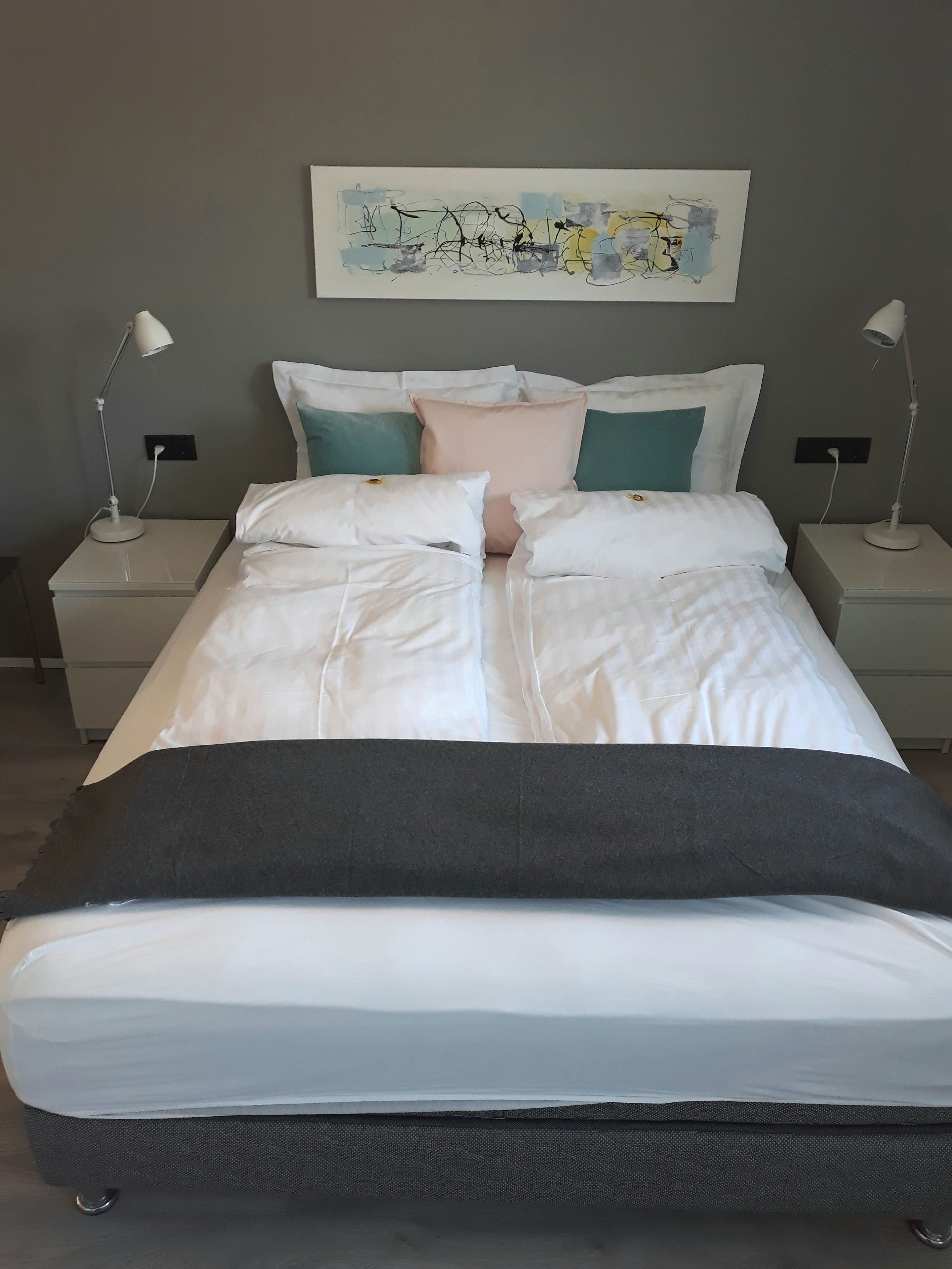 Una cama bien hecha con ropa de cama blanca y negra, dos mesitas de noche con lámparas y una obra de arte abstracto colgada en la pared.