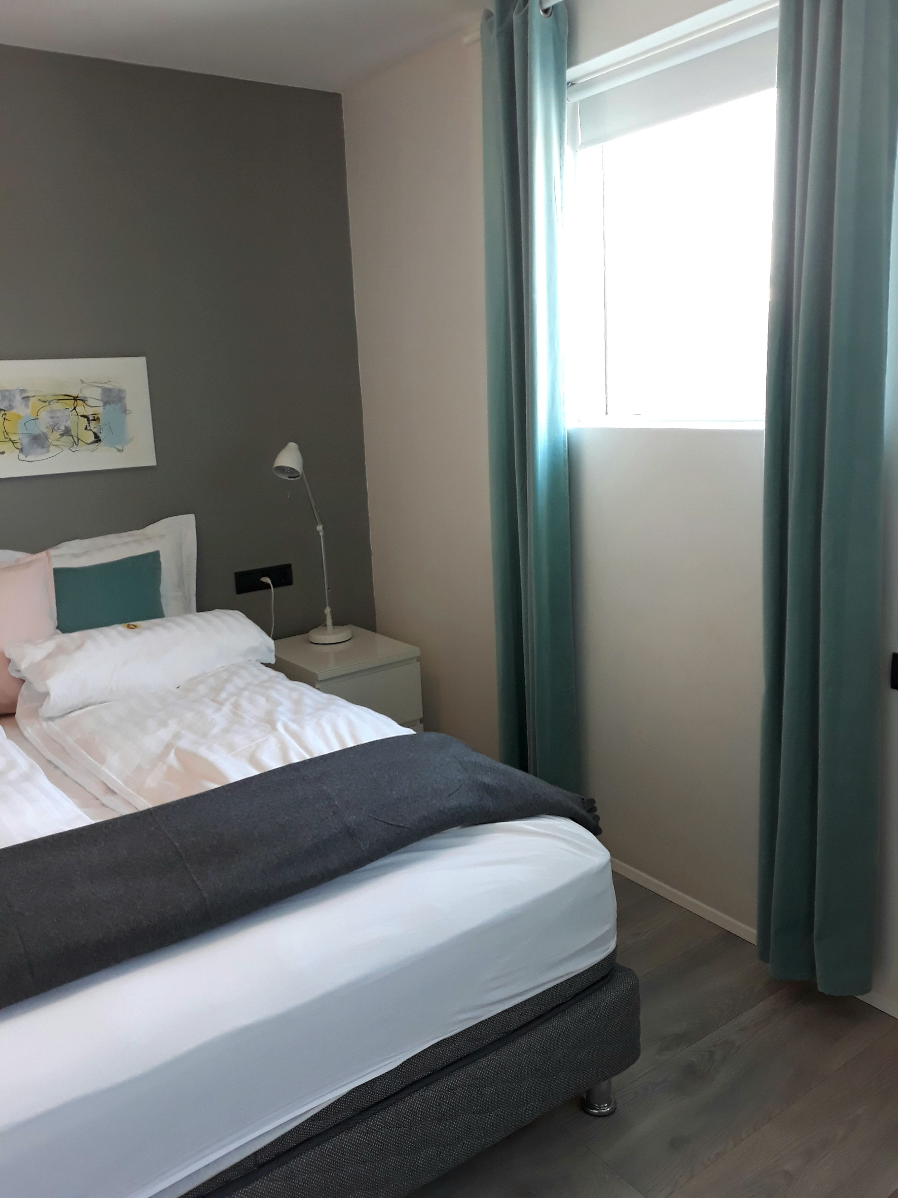 A neatly made bed with white and gray bedding, a small bedside table with a lamp, and a window with curtains.