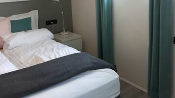 A neatly made bed with white and gray bedding, a small bedside table with a lamp, and a window with curtains.