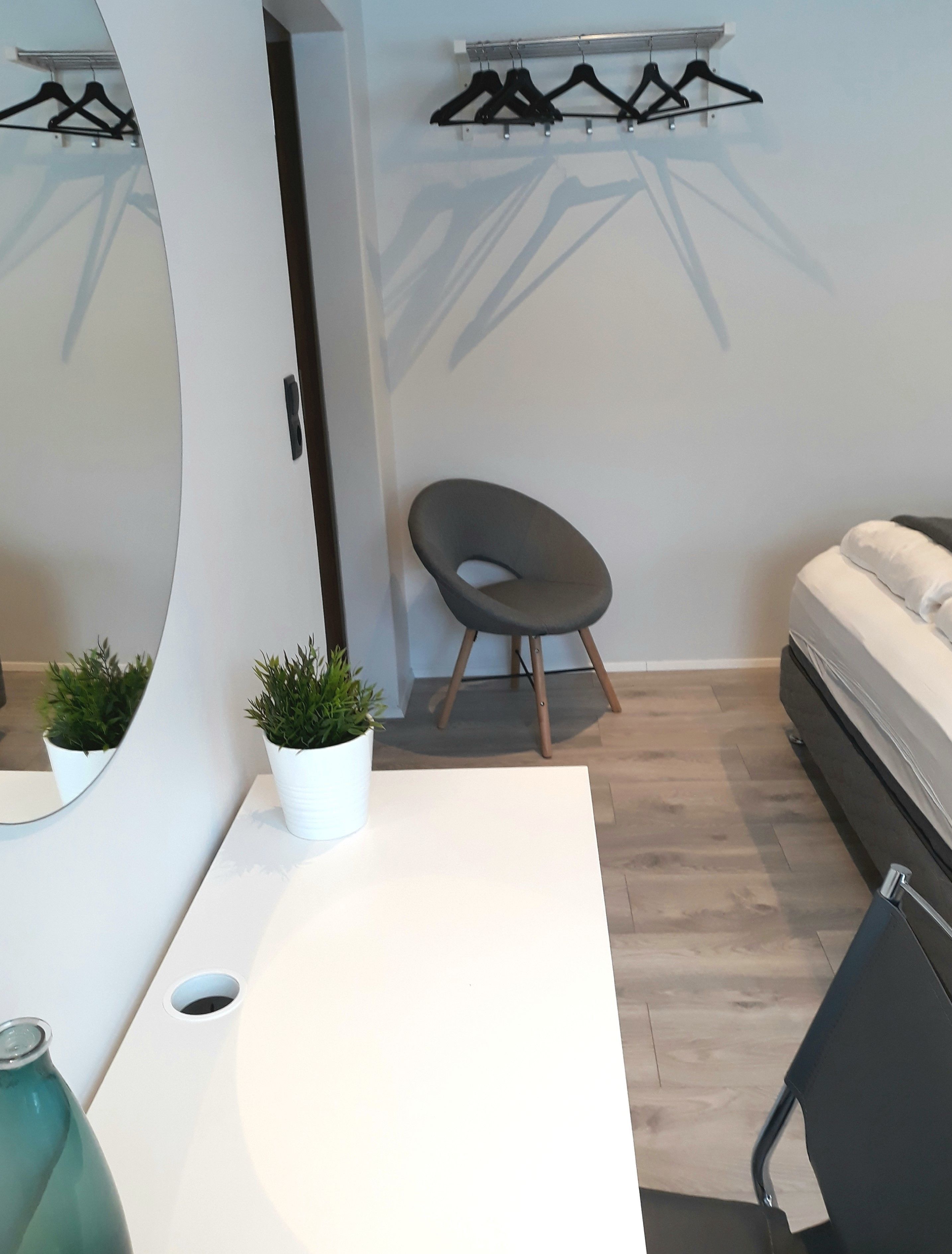 Un dormitorio moderno con una cama blanca, una pequeña mesa redonda con una planta en maceta, una silla y un perchero en la pared.