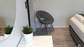 Un dormitorio moderno con una cama blanca, una pequeña mesa redonda con una planta en maceta, una silla y un perchero en la pared.