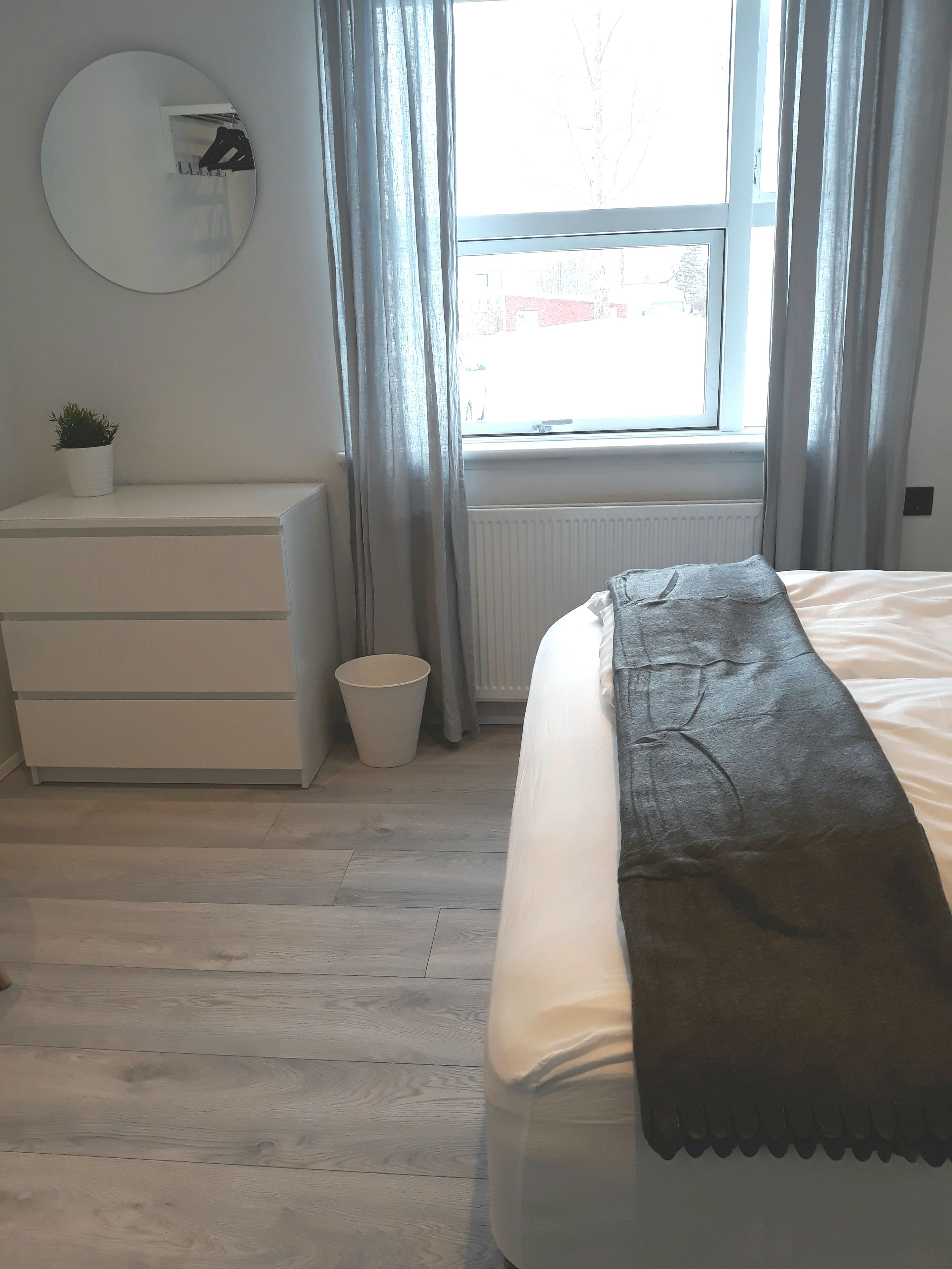 Un dormitorio con cama, cómoda, espejo redondo, ventana con cortinas y radiador.