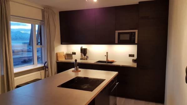 A modern kitchen with a central island, dark cabinetry, and a view of the sea through the window. - Post-Plaza Guesthouse (Husavik)