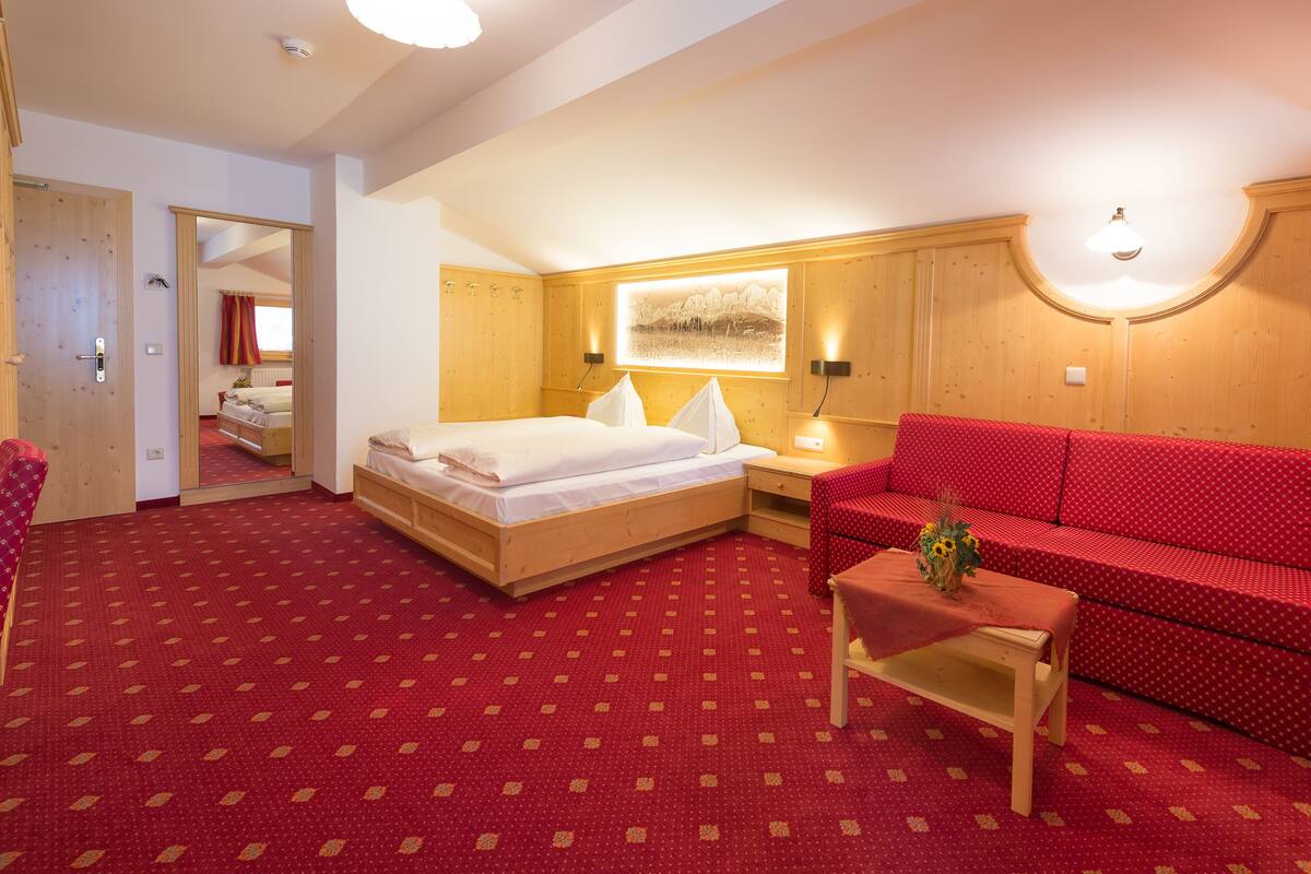 Una habitación de hotel con una cama, un sofá rojo estampado, una mesita con un jarrón y un espejo.