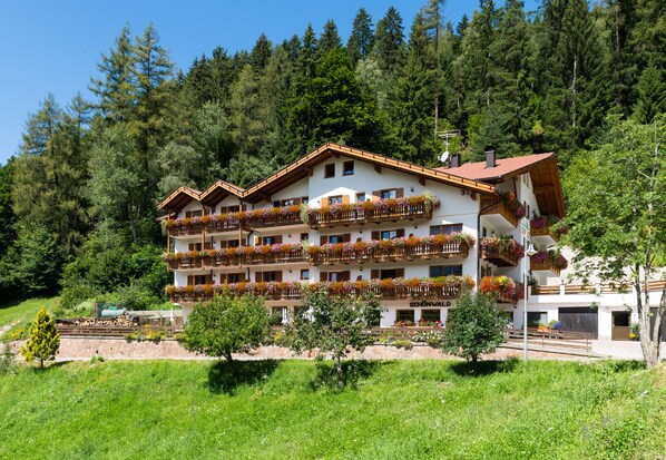 Un hotel de varios pisos con balcones decorados con jardineras de flores, rodeado de abundante vegetación y un fondo de bosque.