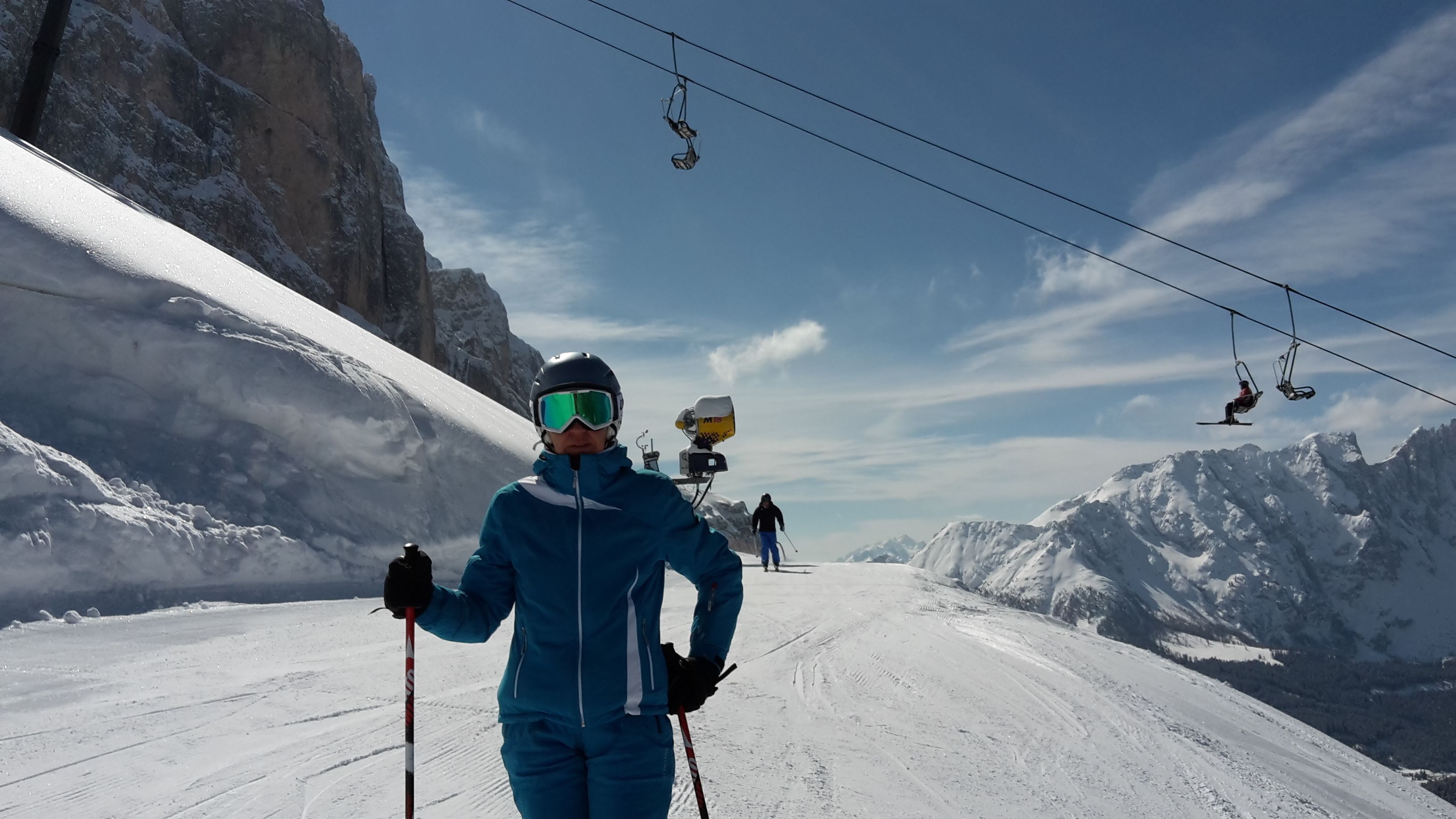 Une piste de ski avec une personne vêtue d’une combinaison de ski bleue et d’un casque, tenant des bâtons de ski, et un télésiège en arrière-plan.