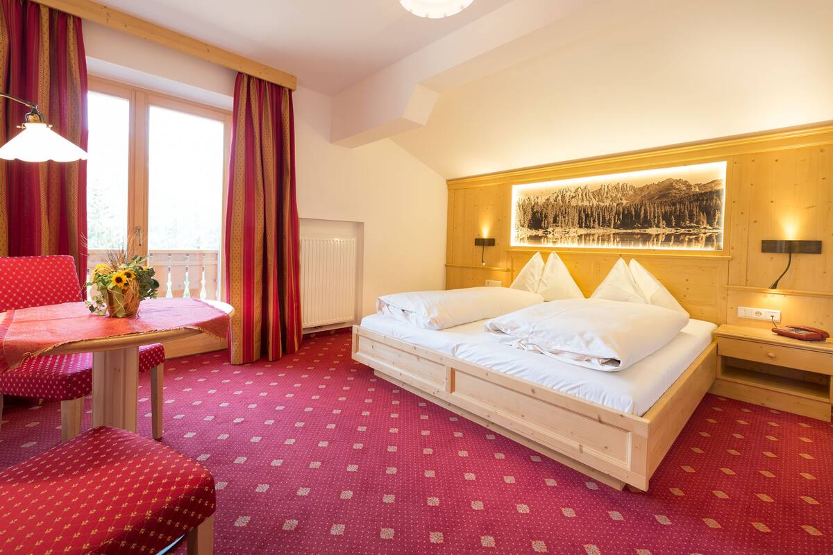 Habitación de hotel con una cama grande, una mesita con una silla, una ventana con cortinas y un cuadro colgado en la pared.
