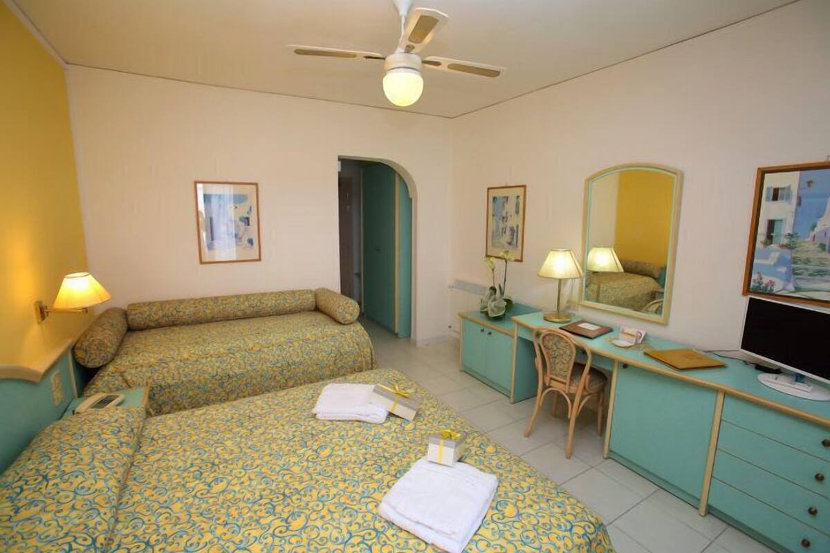 A hotel room with two beds, a desk, a chair, a TV, and a ceiling fan.