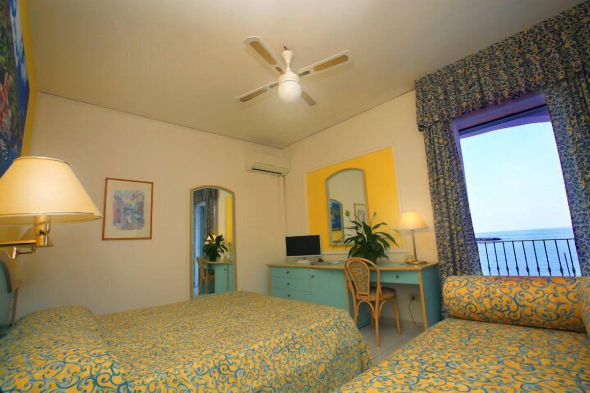 A hotel room with two beds, a desk with a computer, a chair, a ceiling fan, and a window with a view.