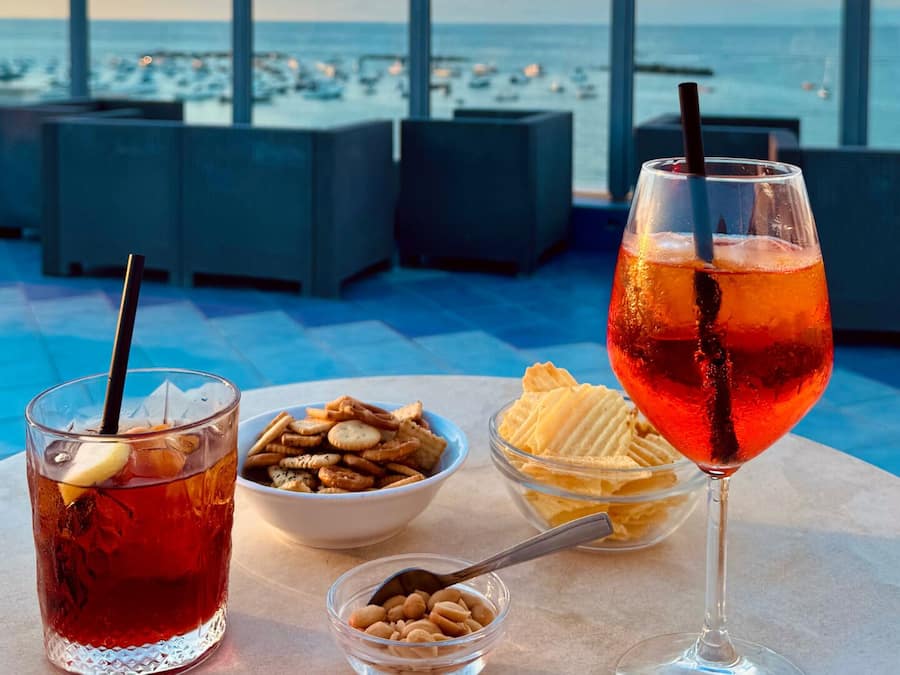Two drinks, a bowl of nuts, and a bowl of ice cream on a table with a view of the sea.