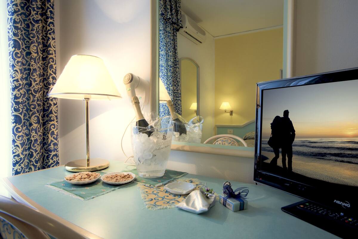 A hotel room with a TV displaying a beach scene, a lamp, a mirror, and a table with various items.