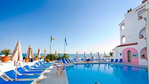 A pool area with lounge chairs and umbrellas, flags, and a building with arches. - Hotel Solemare Beach & Beauty SPA (Ischia)
