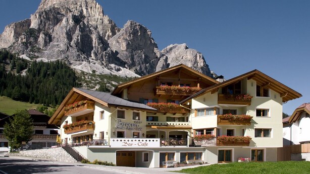 Un edificio de varios pisos con balcones, un letrero que dice 'Bar-Cafe' y un paisaje montañoso de fondo.