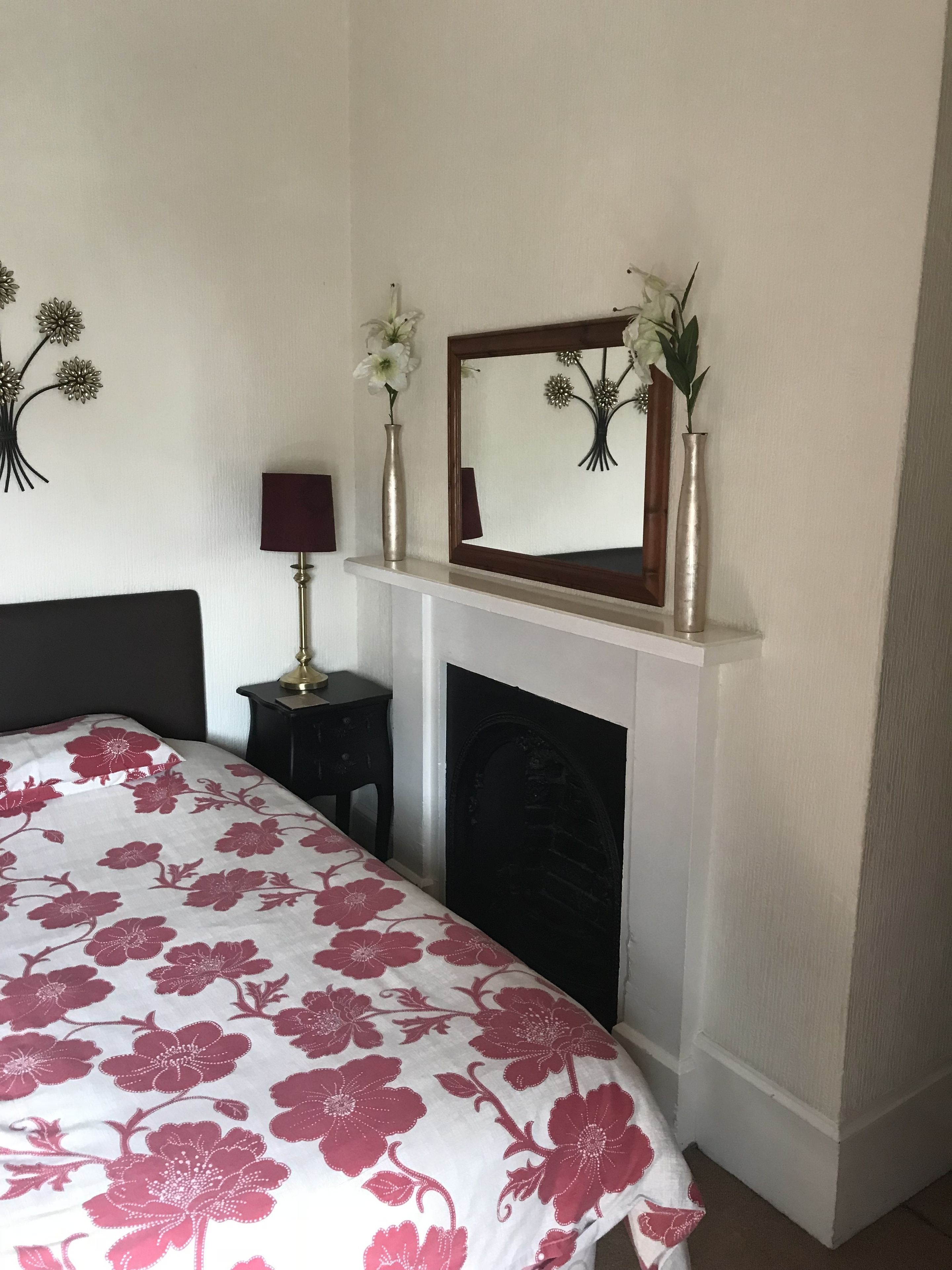 A bedroom with a bed, a fireplace, a mirror, and two vases with flowers.