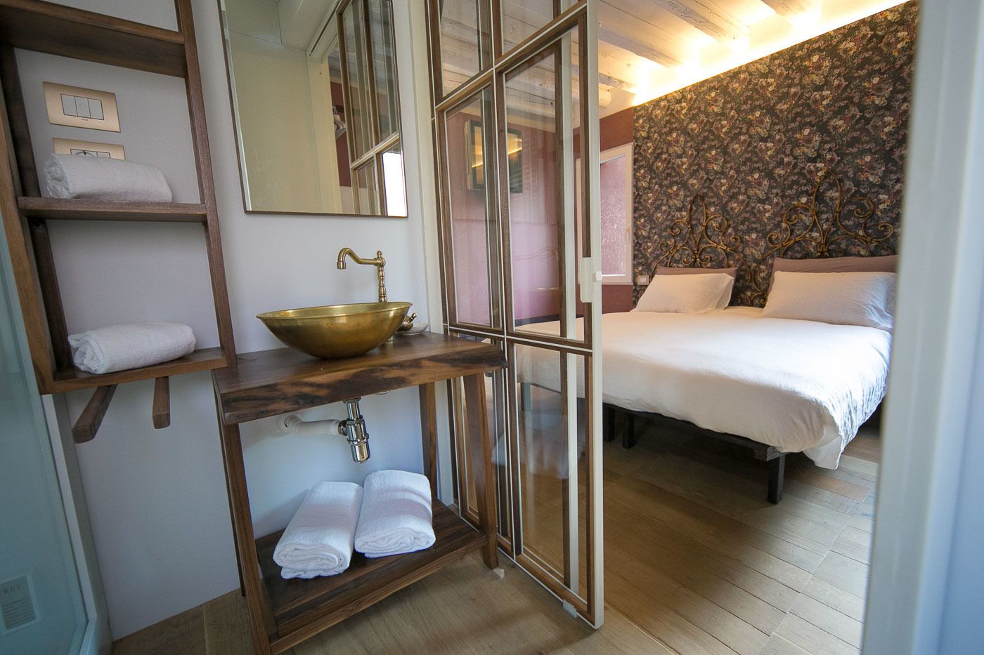 Ein Hotelzimmer mit einem hölzernen Waschtisch, einem Messingbecken, einem Bett mit weißer Bettwäsche und blumigem Wandpapier.