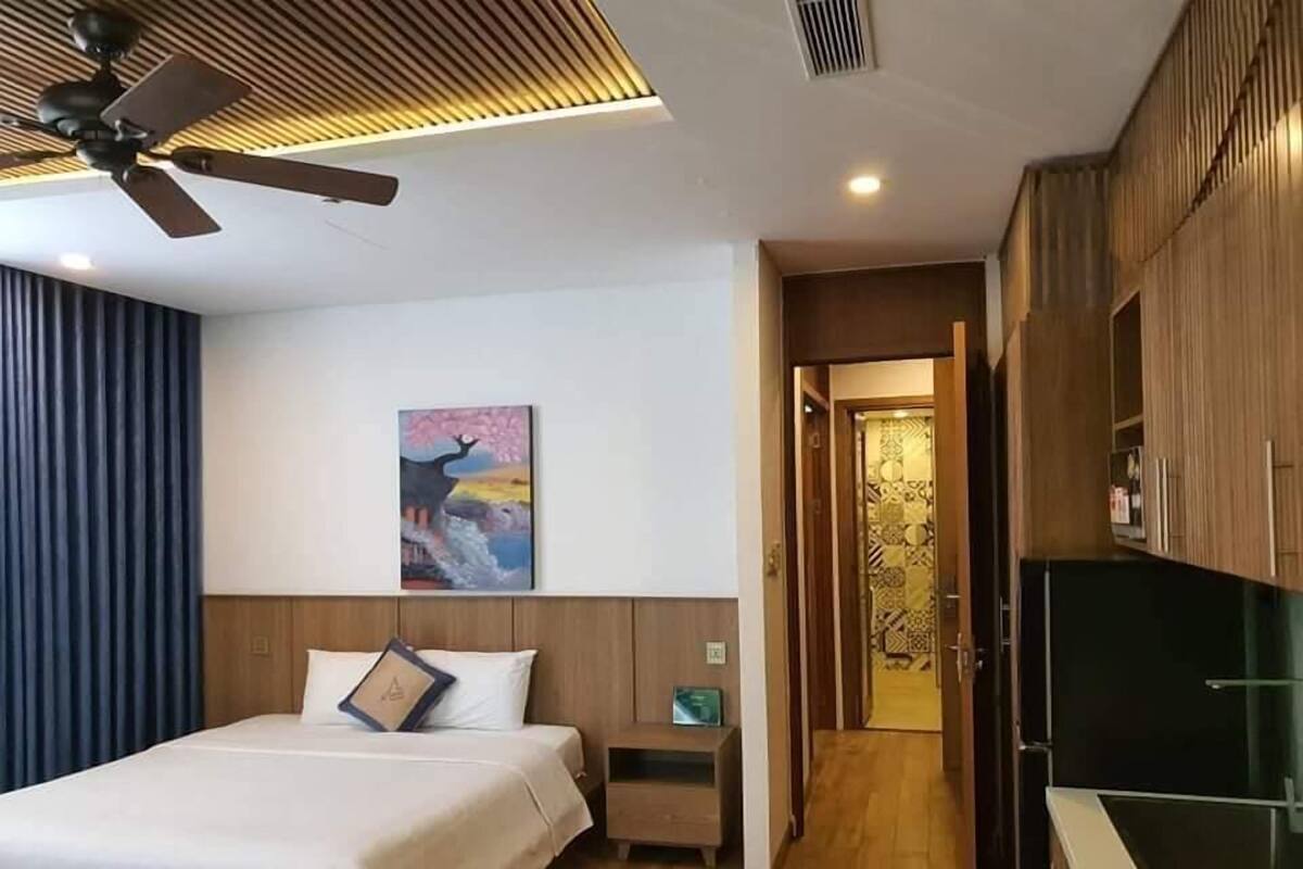 A modern hotel room with a bed, a ceiling fan, a painting on the wall, and a kitchen area.