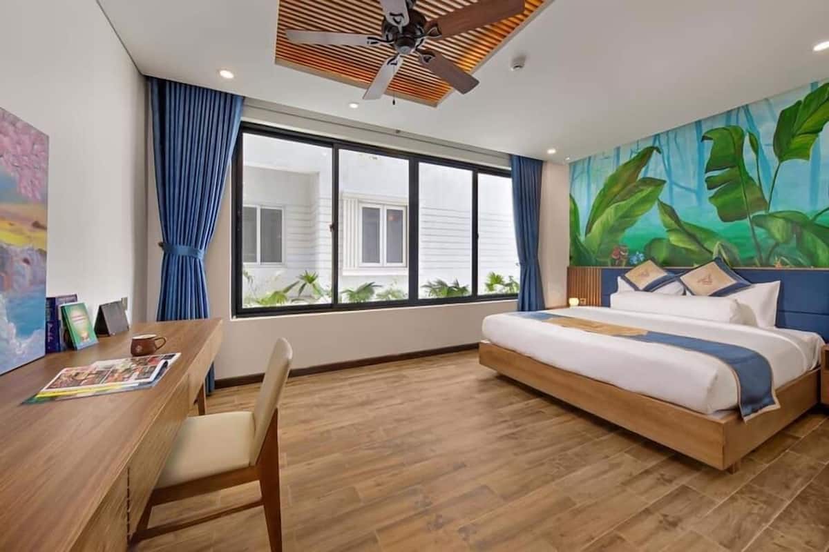 A hotel room with a wooden desk, a bed with a blue headboard, a large window, and a tropical mural on the wall.