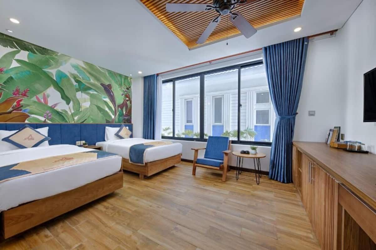 A hotel room with a large bed, a smaller bed, a desk, and a chair. There is a window with curtains, a ceiling fan, and a wall with a tropical mural.