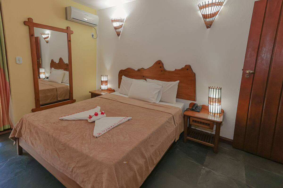 Una camera d'albergo con un letto in legno, un comodino con una lampada, uno specchio e una porta.