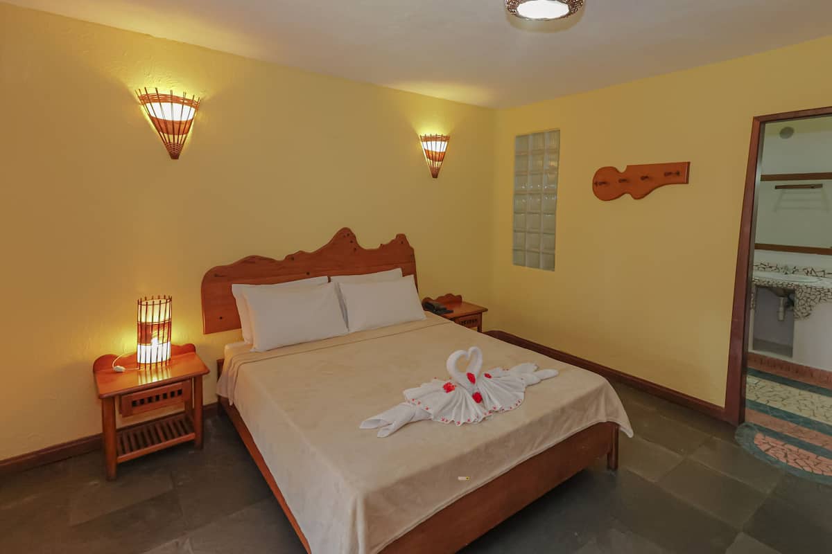Una camera d'albergo con un letto in legno, due comodini con lampade, un punto luce a parete, uno specchio e vista sul bagno.