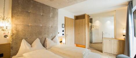 A modern bedroom with a large bed, wooden headboard, and a view into a connected bathroom and kitchen area.
