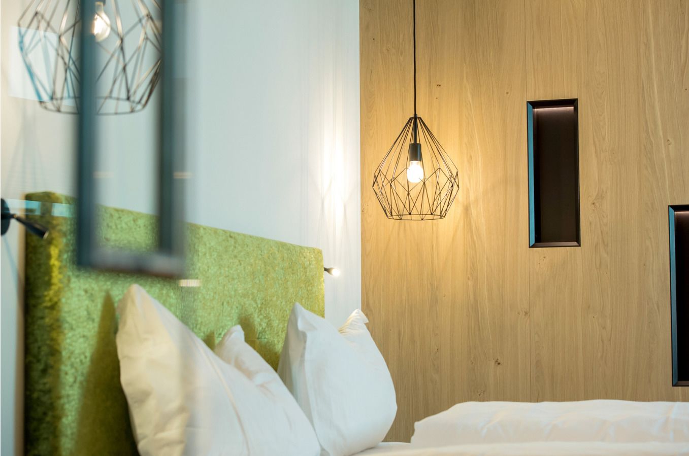 Habitación de hotel con cabecera de madera texturizada en tono verde, lámpara colgante de diseño geométrico y pared de madera.