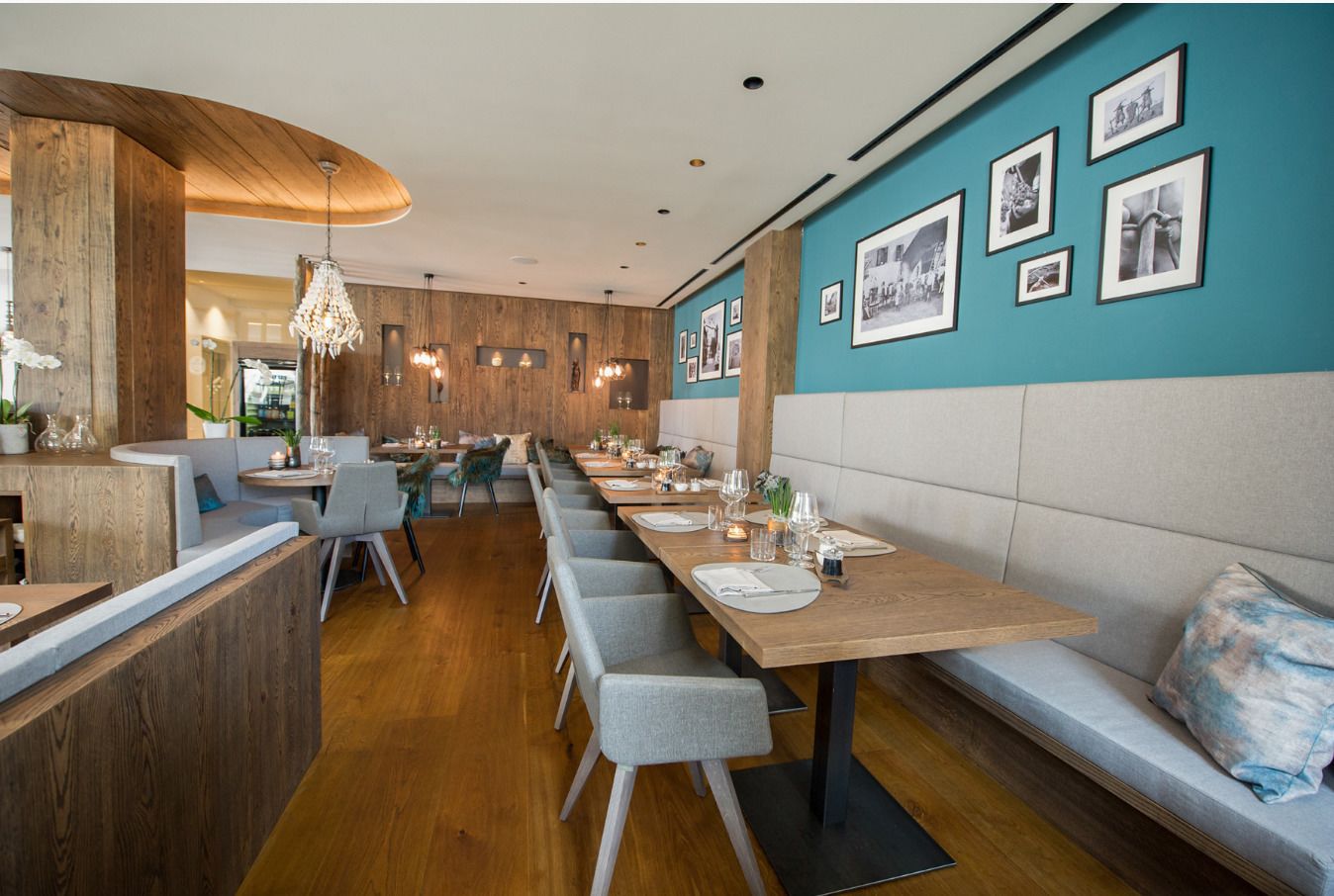 A modern restaurant with wooden accents, grey upholstered seating, and a teal accent wall with framed photos.