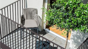 Een balkon met een metalen hekwerk, een rieten stoel met kussen, en een bloempot met plant.