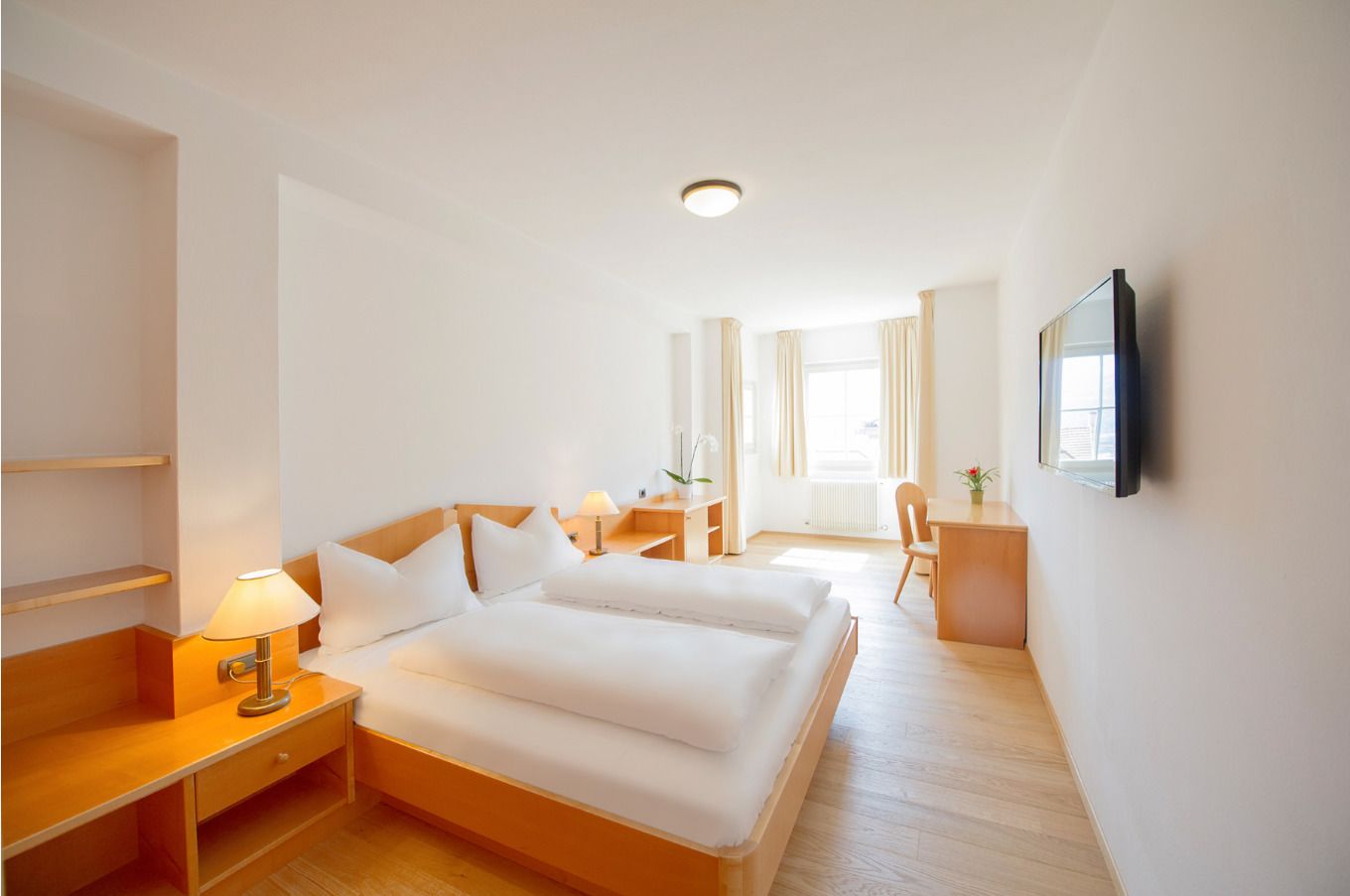 A hotel room with a large bed, bedside tables, a desk, and a TV on the wall.