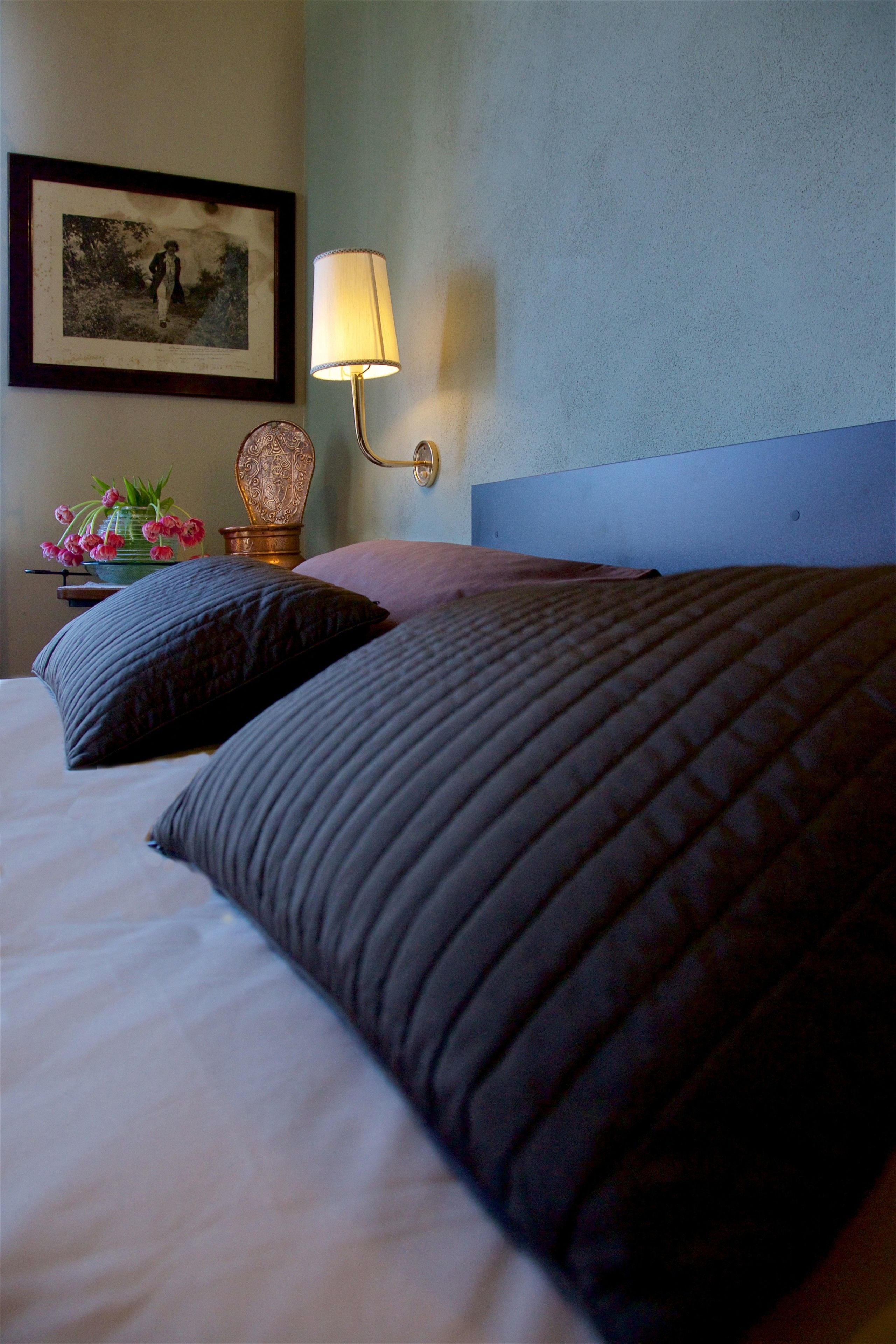 A neatly made bed with dark bedding, a bedside lamp, a framed picture on the wall, and a small table with a vase of flowers.