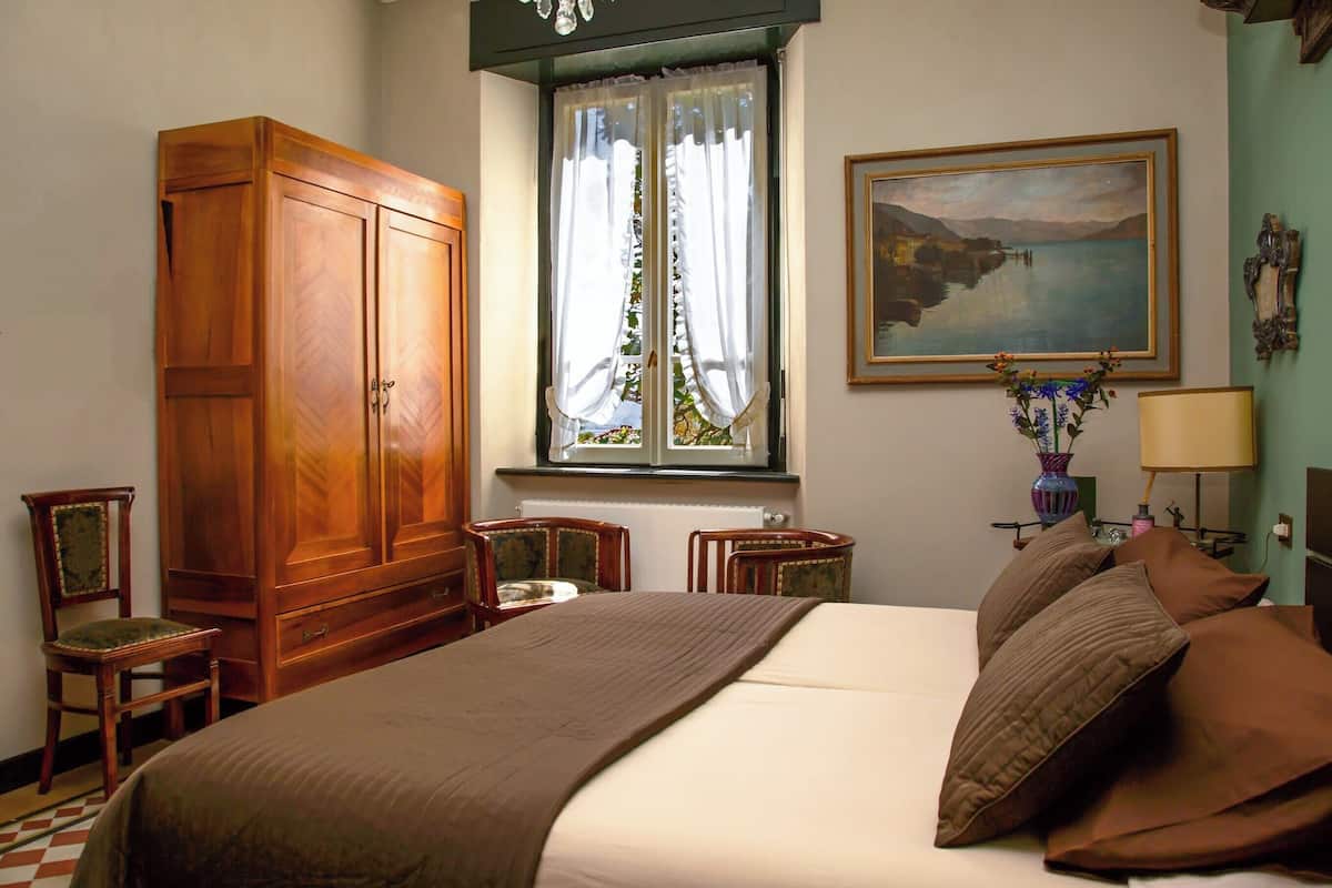 A bedroom with a large wooden wardrobe, a bed with brown bedding, two chairs, a window with curtains, a painting on the wall, and a lamp on a bedside table.
