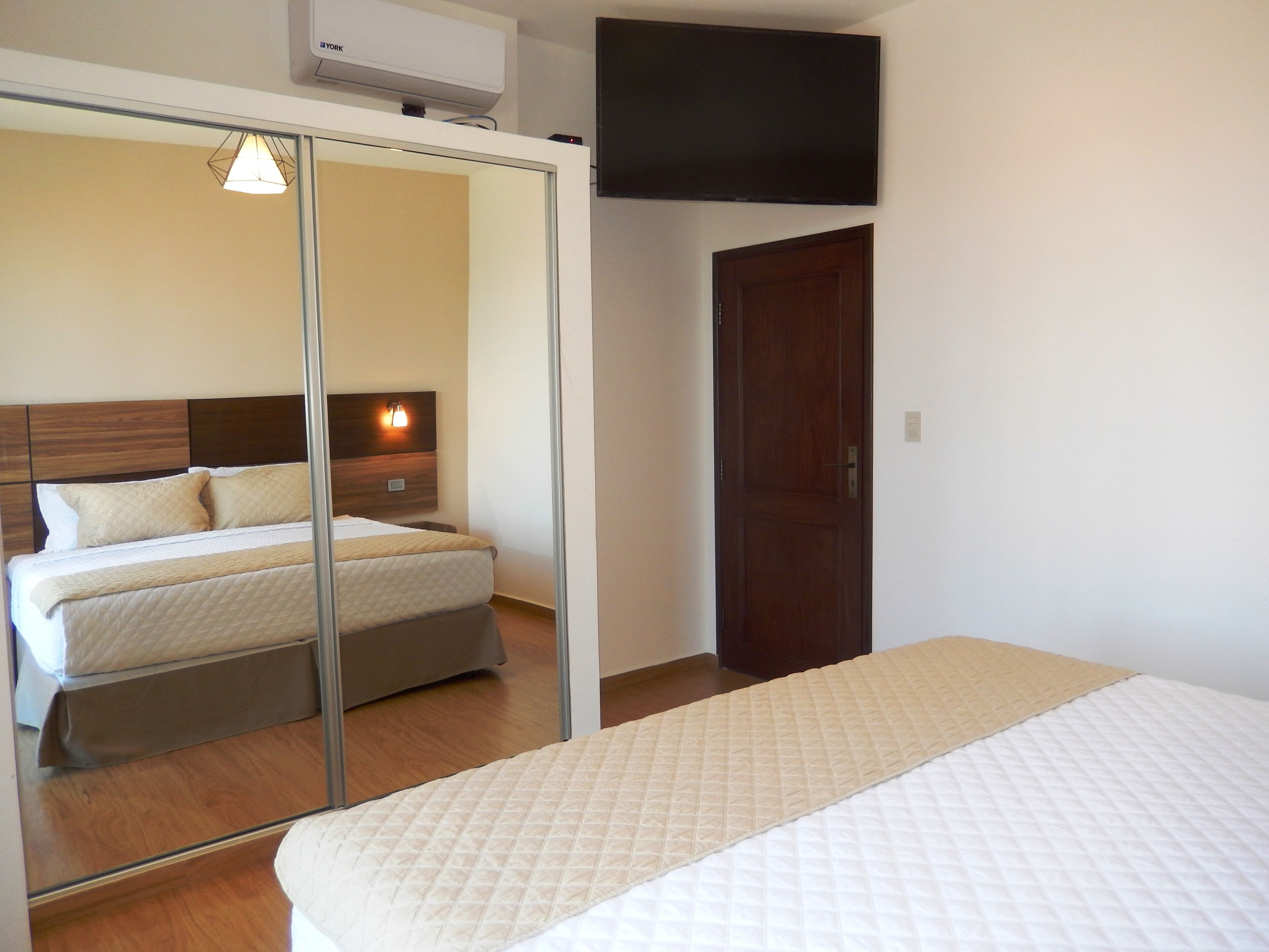 Habitación de hotel con cama, espejo, armario y televisión de pantalla plana montada en la pared.