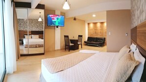 A hotel room with a large bed, a TV mounted on the wall, a dining area with chairs, and a sofa.