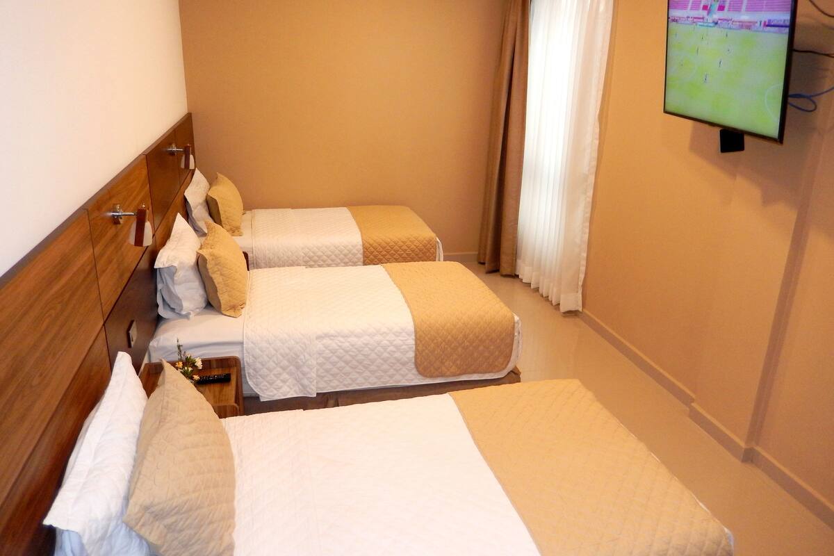 Une chambre d’hôtel avec deux lits, une tête de lit en bois, un téléviseur fixé au mur et une fenêtre avec des rideaux.