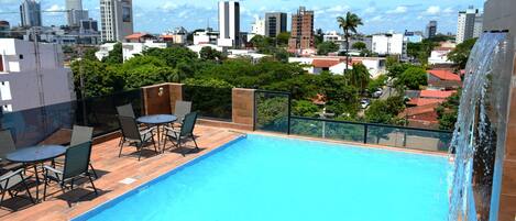 Piscina en la azotea con vista a la ciudad, rodeada de sillones de descanso y una cascada.