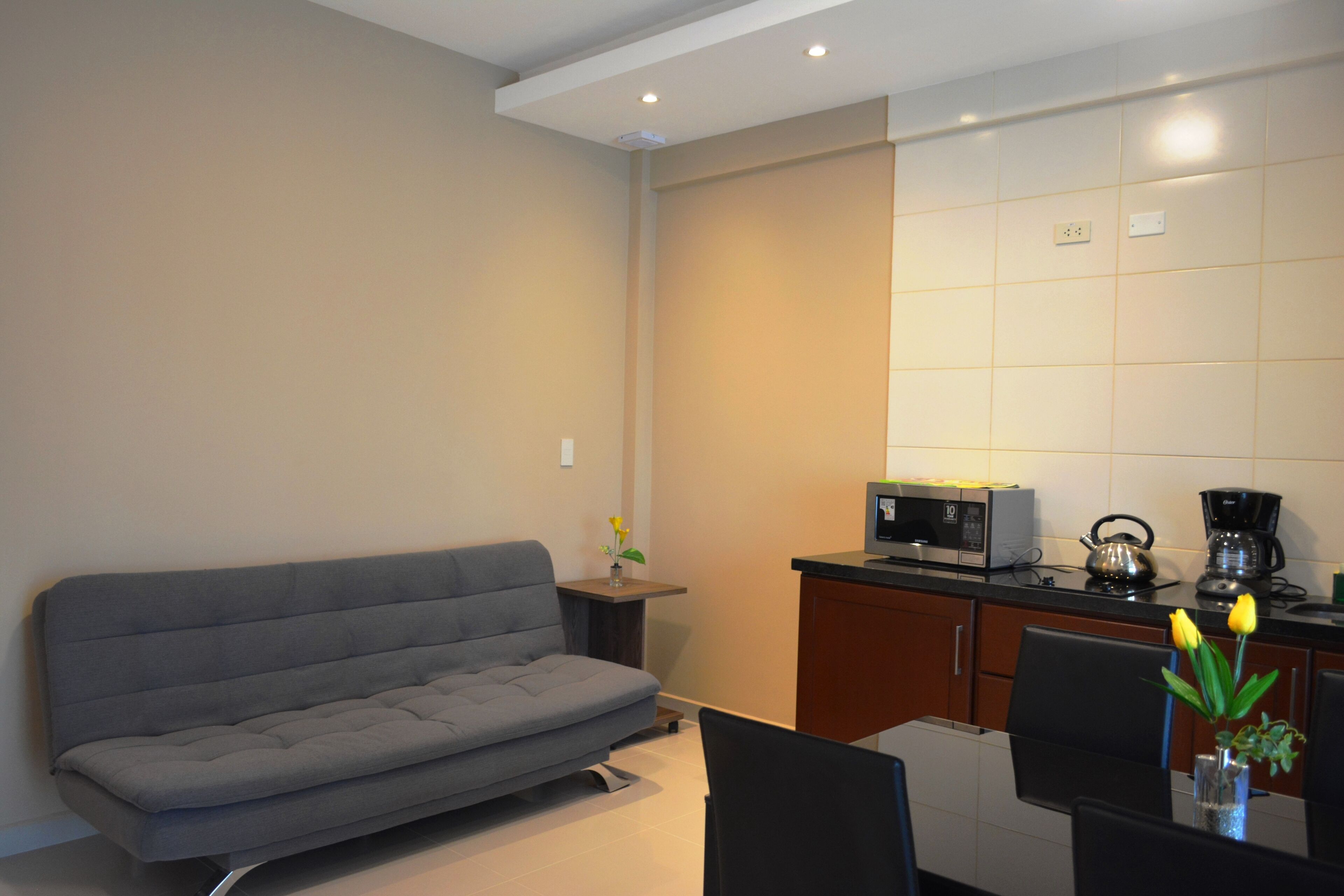 A modern living room with a grey sofa, a dining table with chairs, a small side table with a vase of flowers, and a kitchen area with a microwave, kettle, and coffee maker.