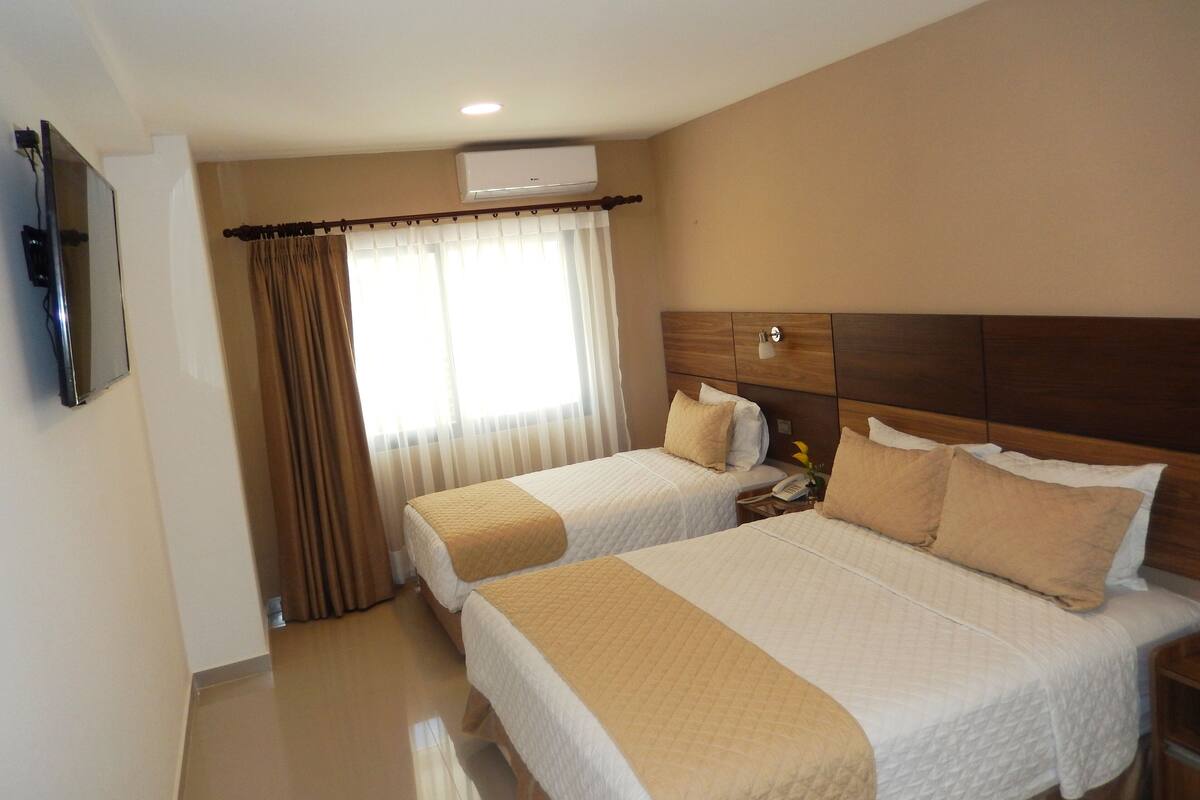 Une chambre d’hôtel avec deux lits, un téléviseur à écran plat fixé au mur, une fenêtre avec des rideaux et un appareil de climatisation placé au-dessus de la fenêtre.
