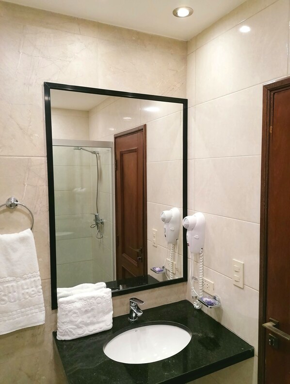 Un baño con un lavamanos negro, un espejo grande, una cabina de ducha de vidrio y un secador de pelo en la pared.